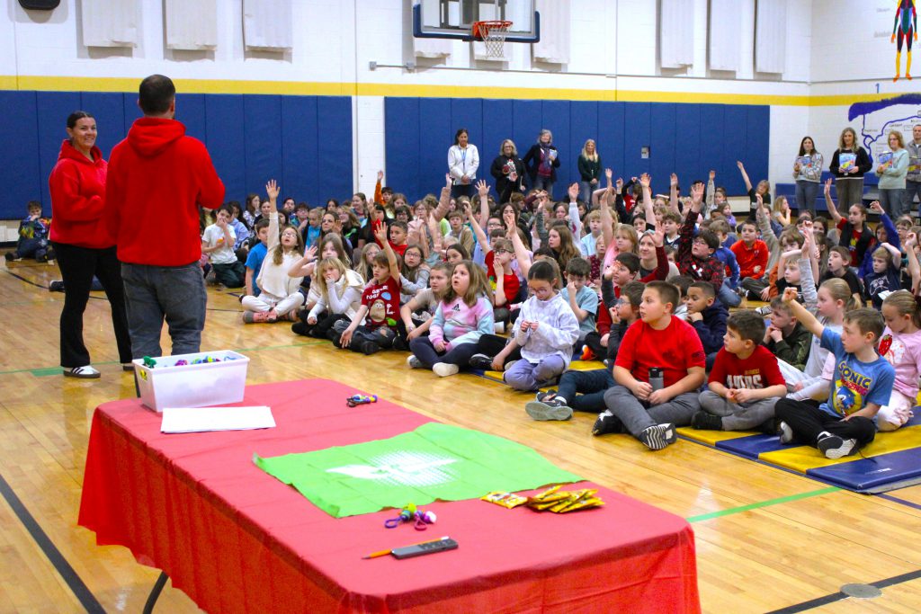 Two teachers wearing red stand before students sitting on the floor with their hands raised