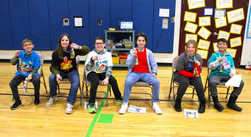 Six students sit in chairs and hold up lanyards with figurines