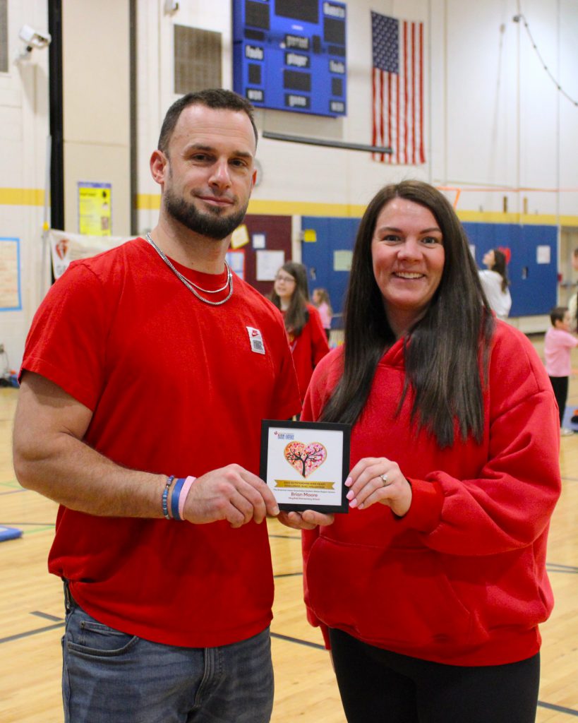 Two gym teachers dressed in red hold a plaque