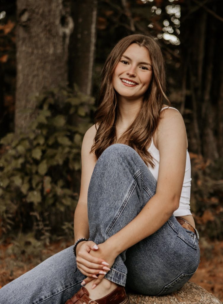student in white sits with knee up in front of a forest background