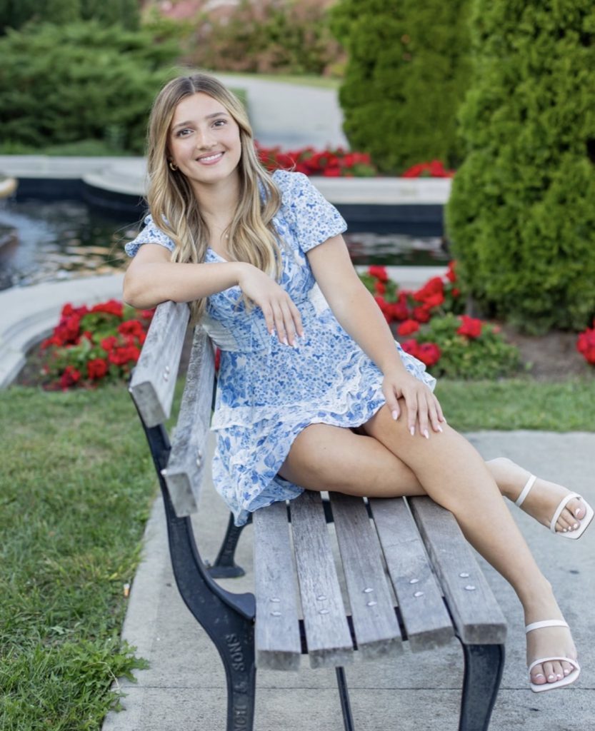 student in blue and white dress on a park bench with red flowers in the background