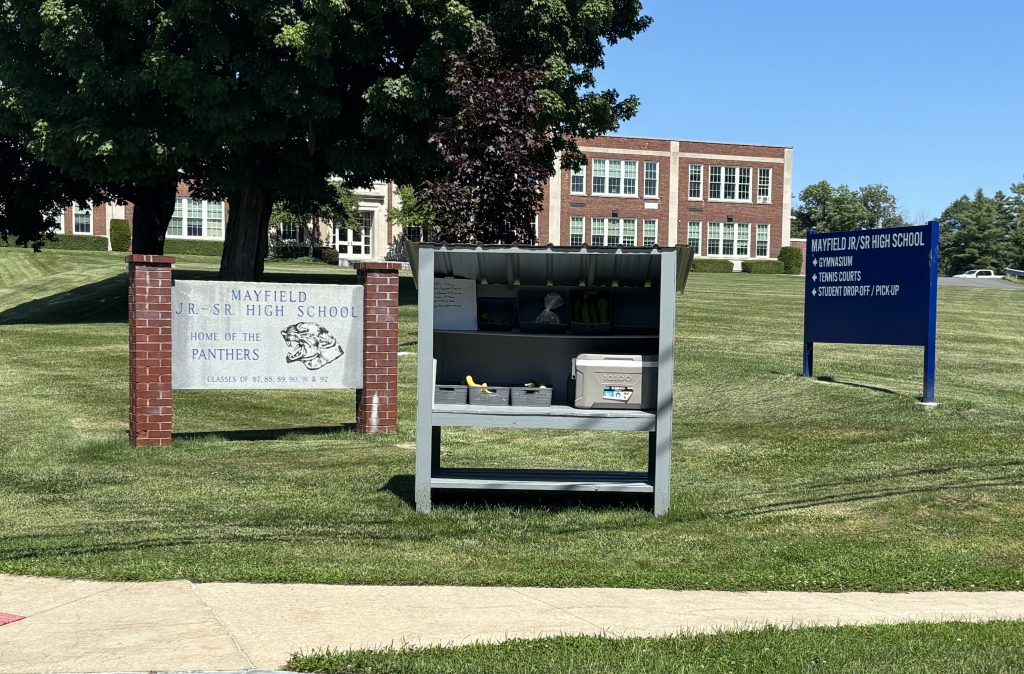 Farm stand on school lawn with signage on either side and brick school building in the background