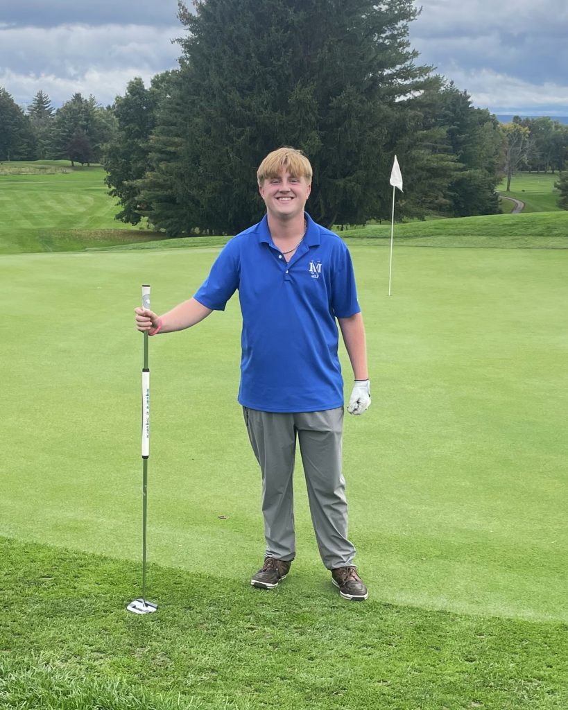Smiling student wearing blue stands on a golf green holding a club