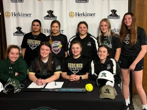 Nine softball players stand in two rows while one player signs a letter. 