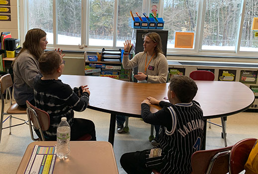 HS student sitting at table talking to elementary students