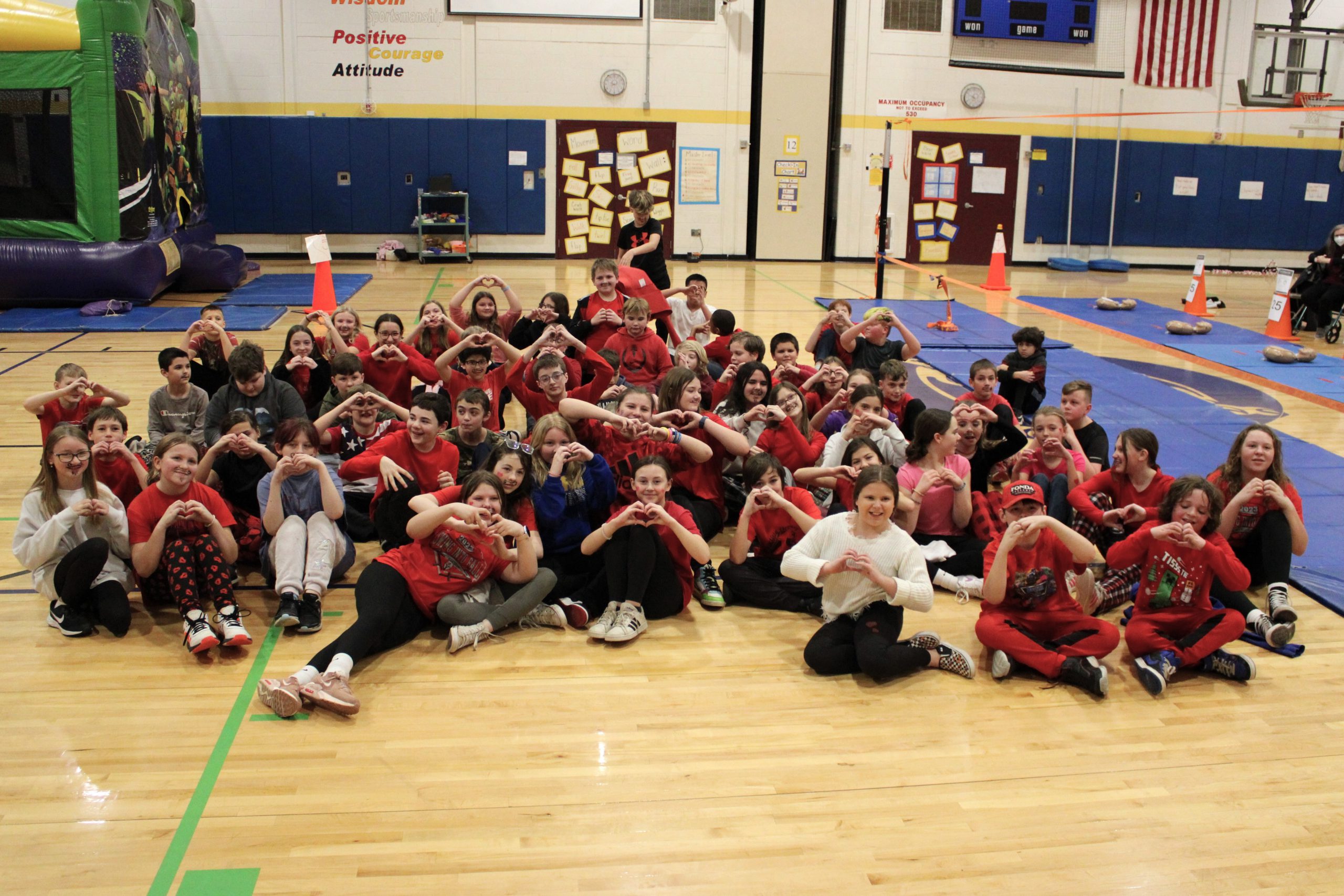 a large group of students wearing red sit on a gym floor and make heart symbols with their hands