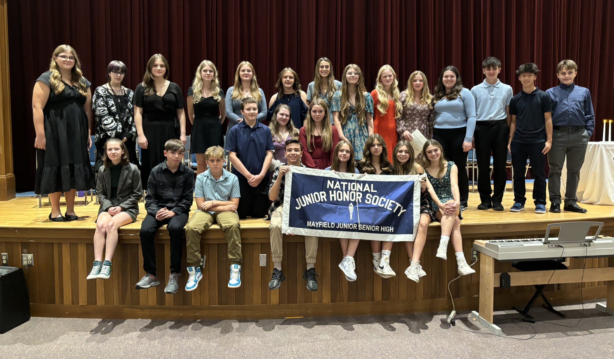 students stand in a row on stage while another row of students sits on the edge of the stage holding a sign saying National Junior Honor Society