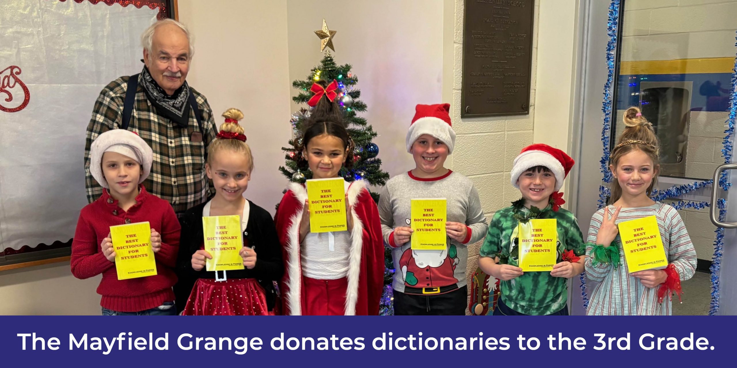 Six students dressed in holiday clothes hold dictionaries. A man in a suspenders and a plaid shirt poses with them. Behind them is a Christmas tree.