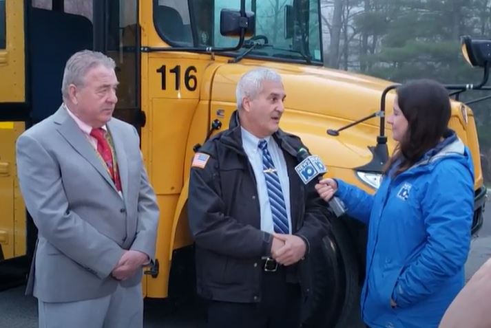 news reporter interviews two men in front of a school bus