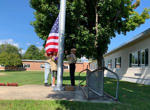 student lowering flag as brother looks on and Scout leader assists